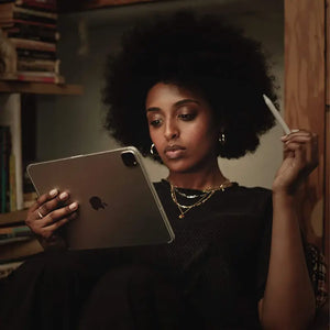 A girl sitting and holding an iPad Pro 2nd gen 11-inch in one hand, watching the screen, while her other hand holds an Apple Pencil. The scene emphasizes her interaction with the iPad and the Pencil in a relaxed, casual setting